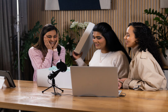Three women smiling, opening white unboxing box at wooden table with laptop, USB mic on tripod