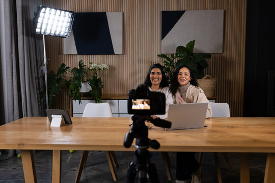 Diverse female co-hosts sitting at wood table in studio filming with camera, LED panel and laptop