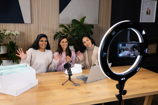 Diverse female hosts in neutral tops smiling, waving to camera in studio with ring light, mic