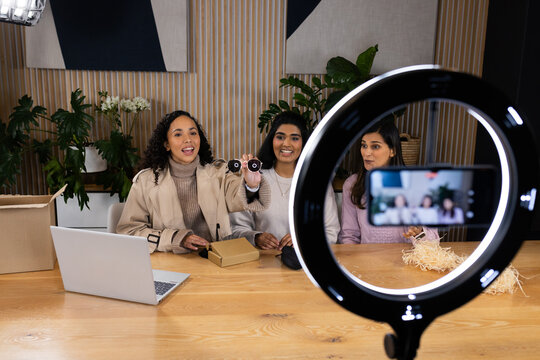 Diverse women hosts in studio, trench, gray, lilac sweaters demonstrating products using ring light