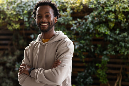 Man posing arms crossed in yard wearing beige hoodie, yellow collar and watch by trellis vines