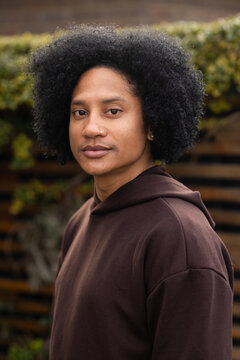 Man standing before wooden slat fence wearing brown hoodie, nose ring and stud earring