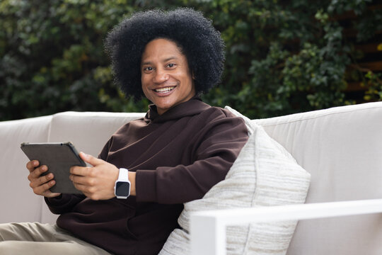 African American man sitting on patio sofa holding tablet with case and wearing smartwatch