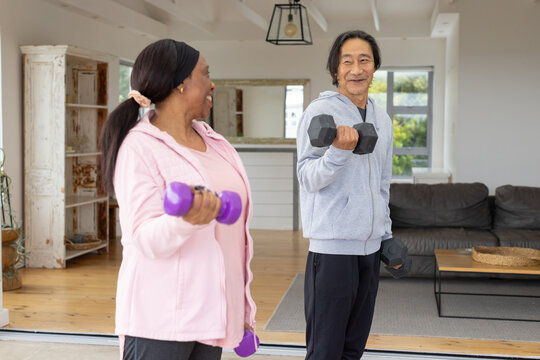 Diverse Senior Asian man and Adult African woman lifting hex and purple dumbbells in living room