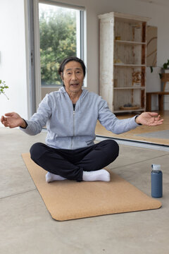 Senior Asian man sitting cross-legged on cork mat at home in hoodie guiding breath with bottle
