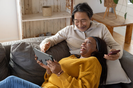 Senior Asian man and African woman sitting on grey sofa at home sharing tablet and smartphone