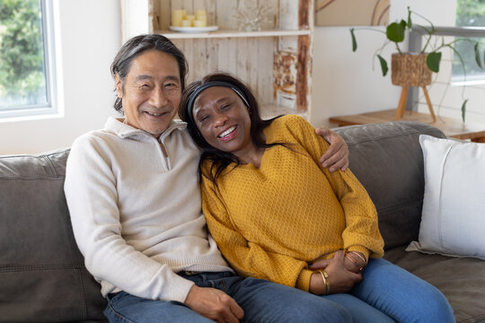 Diverse couple with senior Asian man and woman sitting on gray sofa in living room, smiling