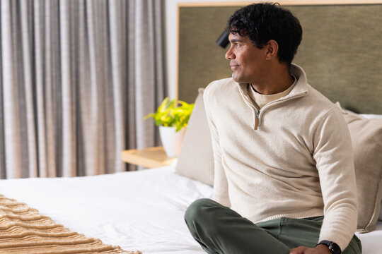 African American man sitting crosslegged on bed facing right with white linen and plant, copy space