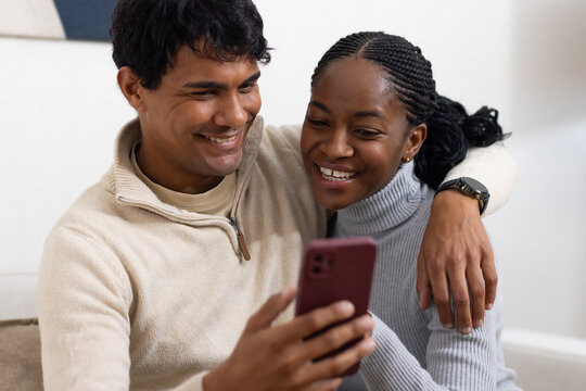 Couple sitting closely on light sofa in living room, viewing smartphone with pink case in sweaters