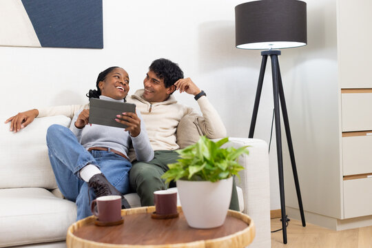 African American couple sitting on sofa in living room holding tablet with coffee mugs