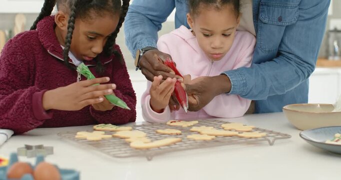 Family decorating sugar cookies for christmas in kitchen, dad guiding girl with red and green bags