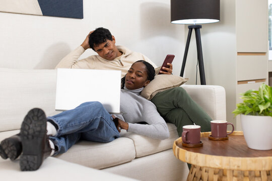 African American couple reclining on light sofa in living room, woman using laptop