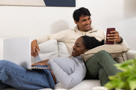 Couple lounging on living room sofa, woman reclining with laptop on lap, man holding smartphone
