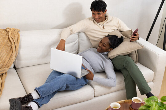 African American couple lounging on sectional sofa at home, woman using laptop, man holding phone