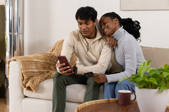 Middle-aged African American couple sitting on sofa at home, checking smartphone and payment card