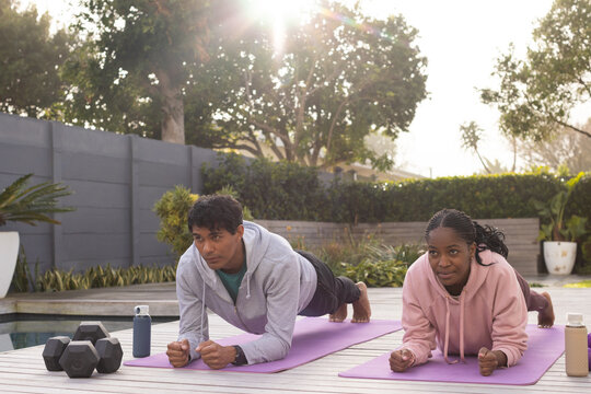 African American friends holding forearm planks on purple yoga mats on deck by pool with dumbbells