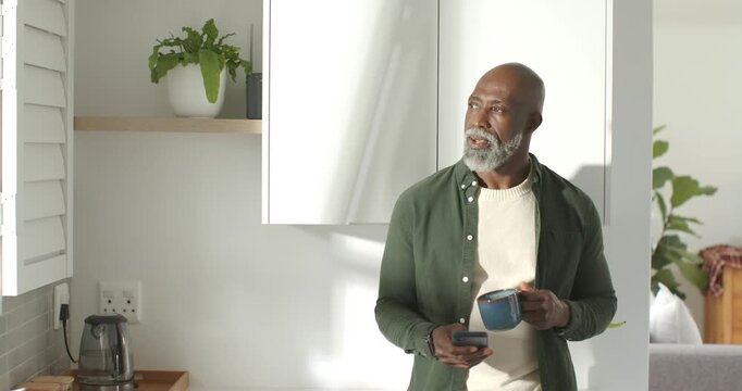 Senior man leaning in kitchen scrolling phone after getting christmas text while sipping mug