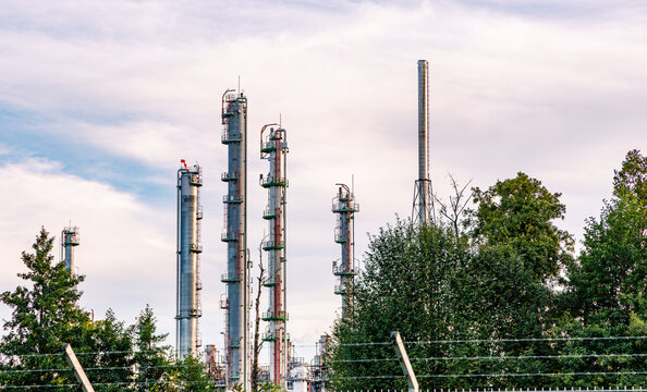 Industrial refinery distillation columns and tall smokestacks rising behind green trees and security boundary fence, chemical plant and energy infrastructure concept