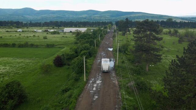 Aerial footage follows a white motorhome driving down a bumpy, wet dirt road through vast green terrain with hills in the background