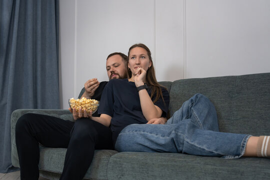 Young couple on a sofa in a modern living room sharing popcorn and watching a movie, relaxed and cozy at home enjoying an intimate, casual evening together