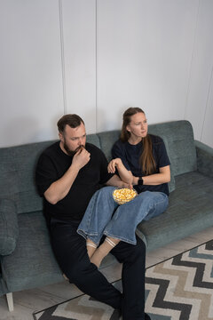Young couple enjoying leisure time together, sitting on the couch in a modern living room, eating popcorn and watching a movie, representing relaxation and comfort. Vertical photo