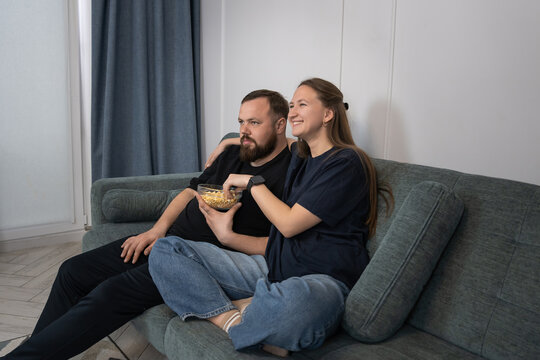 Couple on a sofa in a cozy living room sharing a bowl of popcorn, relaxed and expressive while watching a movie together, enjoying casual home bonding time and comfort
