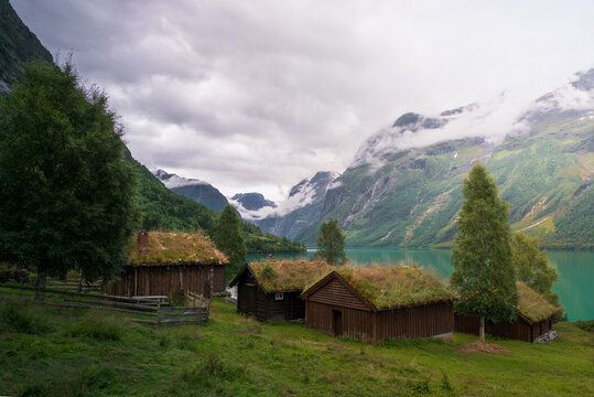 View of traditional wooden cabins with grass roofs situated on the shores of Lovatnet lake, framed by dramatic mountains and a cloudy sky in Stryn Municipality, Vestland, Norway.