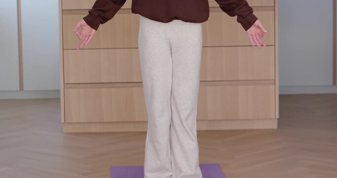 Frame revealing female raising arms above head for breathing on purple mat before dresser in hoodie