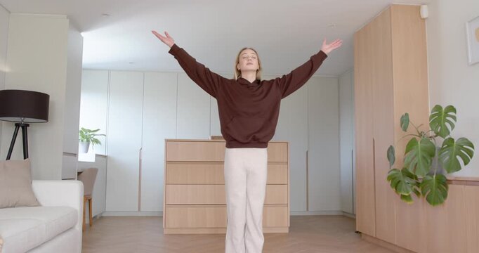 20s woman starting arms outstretched, lifting overhead on purple mat at home, brown top for stretch