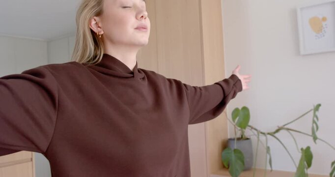 Woman taking deep breath, lifting arms, joining palms for calm in living room near plant cabinet