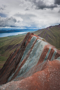 View of the vibrant, multi-colored mineral layers of Pallay Punchu Rainbow Mountain overlooking a vast lake under a dramatic cloudy sky in Layo, Cuzco, Peru.