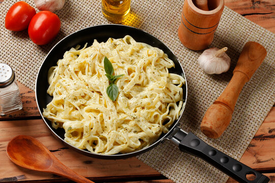 Fettuccine Alfredo with creamy cheese sauce in a black skillet, accompanied by basil leaves on a rustic wooden table.