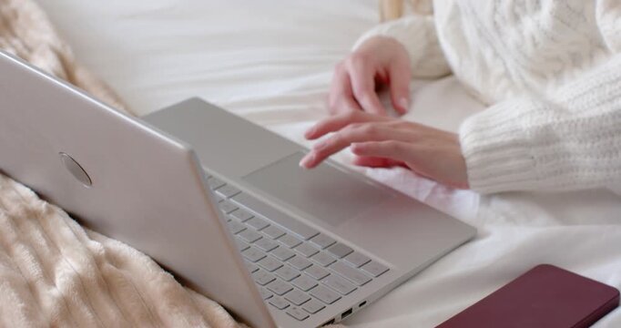 Woman in 20s typing on laptop on bed wearing cream sweater, red smartphone, smiling at message