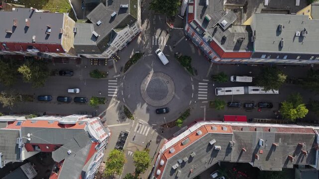 Bird's eye view of a roundabout on a tree-lined street with rows of parked cars in Hamburg, Germany, as an articulated bus turns out, red awnings and long shadows