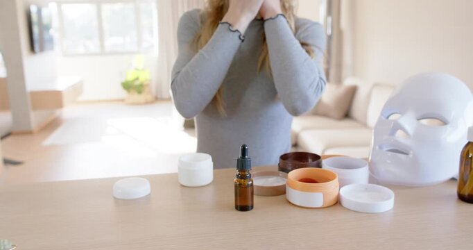 Woman bringing hands to face, using green gua sha massaging jawline at wooden counter for skincare