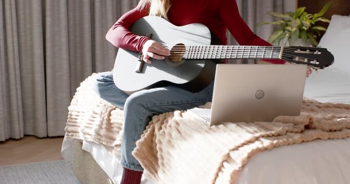 Prompted by laptop chord chart, woman sitting on bed in red top strumming guitar practicing song