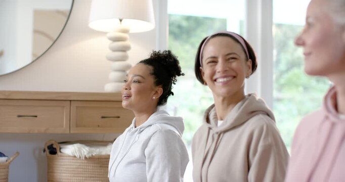 Three women friends smiling and leaning in hoodies in living room, responding to off-screen speaker