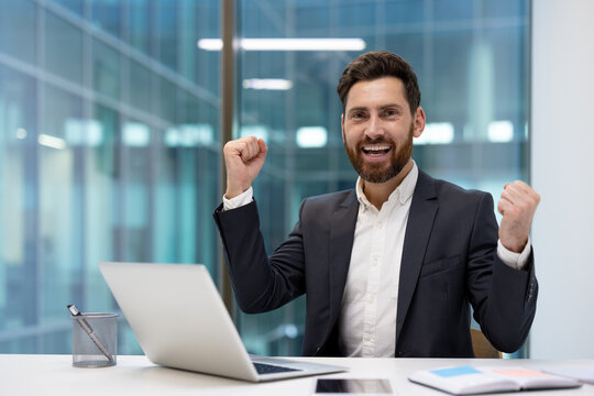 A happy young businessman sits at his office desk with a laptop, smiling at the camera, raising his hands in the air, and celebrating