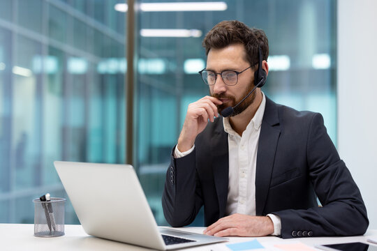 A photo of a thoughtful young businessman sitting at an office desk wearing a headset and looking thoughtfully at a laptop screen