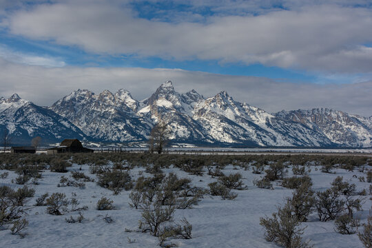 View of the snow-capped Teton Range rising above the sagebrush-covered valley floor and a historic wooden barn in Jackson Hole, Teton County, Wyoming, United States.