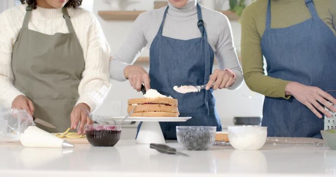 Diverse women helping center baker layering and icing cake finishing decor with spatula in kitchen