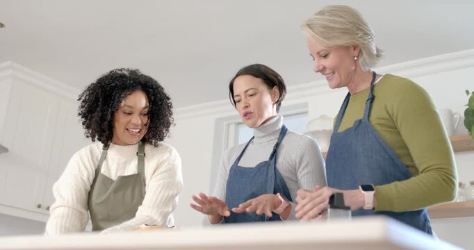 Diverse women in aprons spotting cake at kitchen island, checking layers and smoothing with scraper