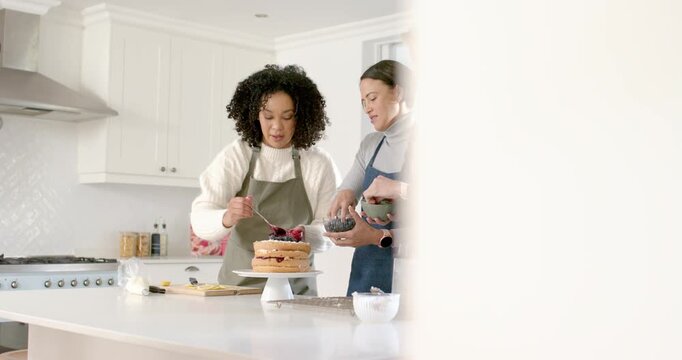 Diverse women in aprons in kitchen left woman spooning fruit joining arranging layer cake to serve
