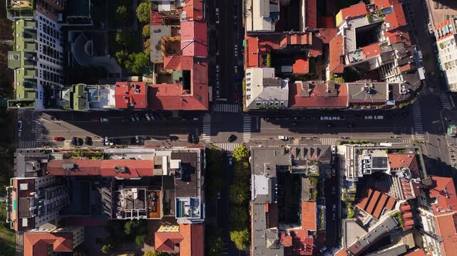 Top down flight over red tiled courtyard blocks and crosswalks toward a pedestrian square with pastel checkered paving and a round building in low sunlight, Milan, Italy
