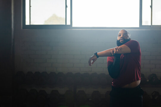 African American man stretching left-arm at gym by dumbbells, maroon-top wrist-tracker, copy space
