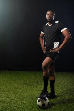 African American man standing on artificial turf, foot on soccer ball, wearing black kit