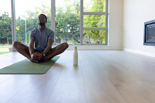 African American man stretching in butterfly pose on green mat at home, water bottle nearby