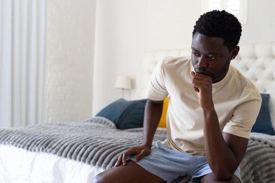 African American man sitting on bed in bedroom by tufted headboard, wearing beige tee, copy space