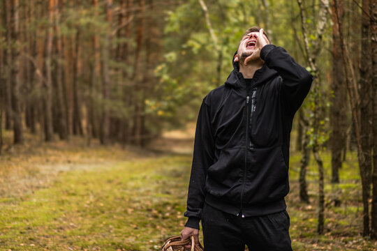 Man in black hooded clothing calling out while carrying a wicker basket along a forest path surrounded by tall trees and mossy ground