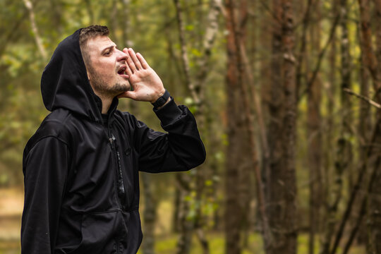 Man in black hooded jacket calling out with cupped hands among tall trees in a sunlit forest, outdoor portrait with natural background